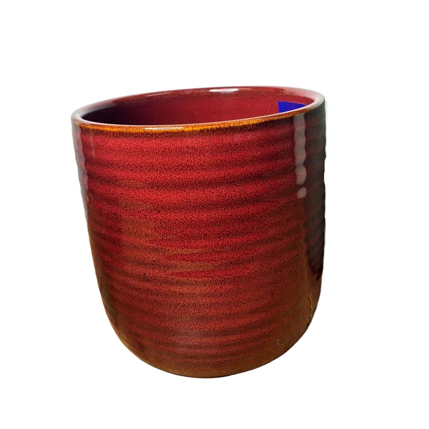 A handmade ceramic cup with a textured, ribbed exterior and a glossy deep red-brown glaze, sitting on a wooden table next to another ceramic planter.